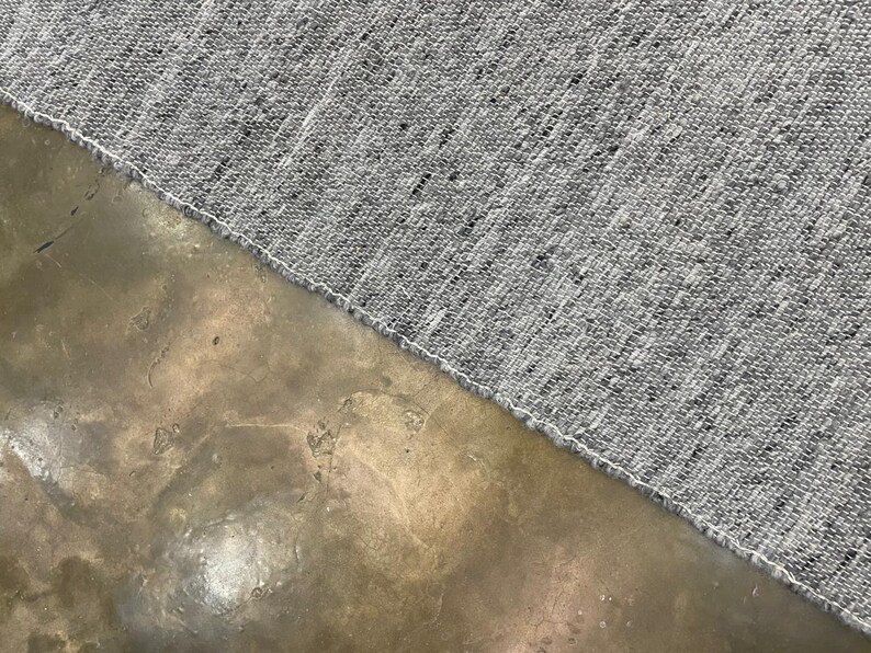 May include: Close-up of a grey textile with a textured weave, featuring flecks of black and white. The fabric's edge is finished with a white seam. The textile is set against a mottled, brown surface.