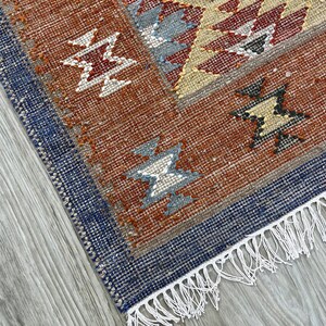Könnte beinhalten: Nahaufnahme eines gewebten Teppichs mit geometrischem Muster. Der Teppich hat eine Farbpalette aus Rost, Blau, Beige und Grau. Das Design umfasst sternartige Formen und einen Fransenrand. Der Teppich liegt auf einem hellgrauen Holzboden.