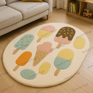 May include: An oval-shaped rug with a cream-colored background features colorful ice cream designs. The rug has various ice cream popsicles and ice cream cones in shades of pink, yellow, orange, and blue. The rug is made of a soft, plush material.