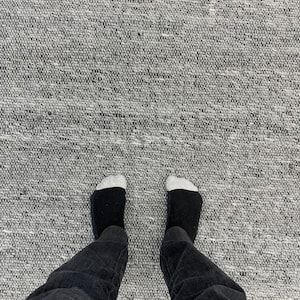 May include: A person's legs and feet are visible, wearing black jeans and black socks with white toes, standing on a grey woven rug. The rug has a textured appearance.