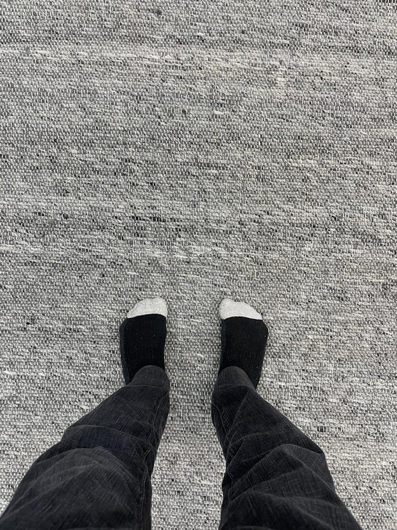 May include: A person's legs and feet are visible, wearing black jeans and black socks with white toes, standing on a grey woven rug. The rug has a textured appearance.
