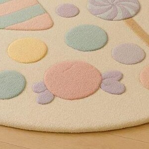 Puede incluir: Una alfombra redonda de color crema con dise&ntilde;os de caramelos en tonos pastel. La alfombra presenta varias formas, incluyendo piruletas, caramelos y formas rayadas en tonos rosa, morado, amarillo y verde. La alfombra est&aacute; colocada sobre un suelo de madera clara.