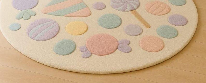 Puede incluir: Una alfombra redonda de color crema con dise&ntilde;os de caramelos en tonos pastel. La alfombra presenta varias formas, incluyendo piruletas, caramelos y formas rayadas en tonos rosa, morado, amarillo y verde. La alfombra est&aacute; colocada sobre un suelo de madera clara.