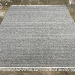May include: A rectangular grey rug with a textured surface and a white fringe along the bottom edge. The rug is made of a woven material. The rug is placed on a concrete floor. The rug measures approximately 240 cm x 300 cm.