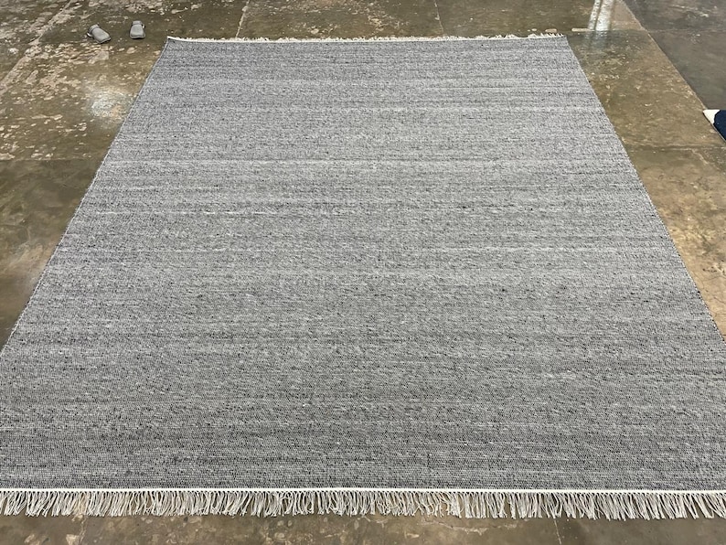 May include: A rectangular grey rug with a textured surface and a white fringe along the bottom edge. The rug is made of a woven material. The rug is placed on a concrete floor. The rug measures approximately 240 cm x 300 cm.