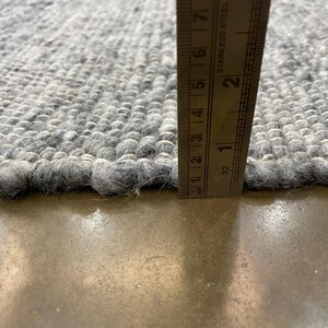 May include: Close-up of a grey rug with a textured, woven design. A stainless steel ruler shows the rug's thickness, approximately 2.5 cm. The rug's edges are slightly frayed. The rug is on a concrete surface.