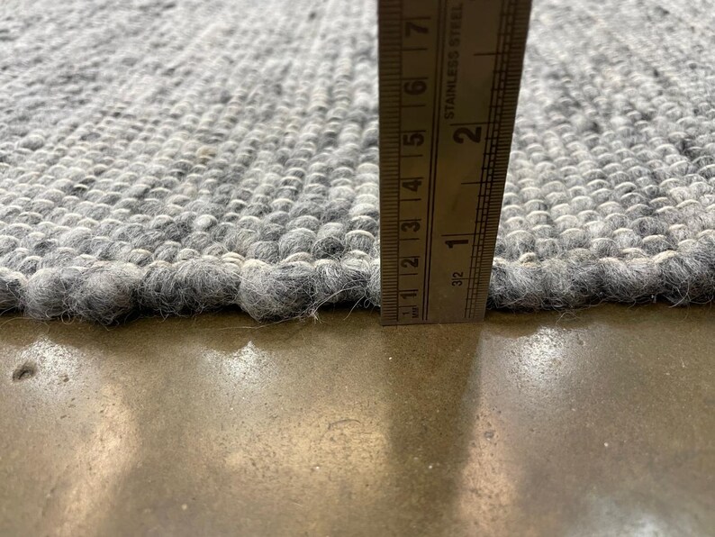 May include: Close-up of a grey rug with a textured, woven design. A stainless steel ruler shows the rug's thickness, approximately 2.5 cm. The rug's edges are slightly frayed. The rug is on a concrete surface.