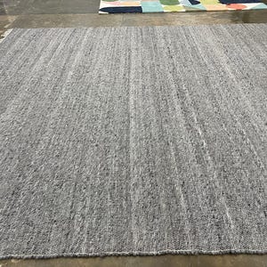 May include: A large, rectangular grey rug with a textured surface. The rug has a woven appearance with visible vertical lines and a fringe along the edge. The neutral colour and simple design make it suitable for various interior styles.