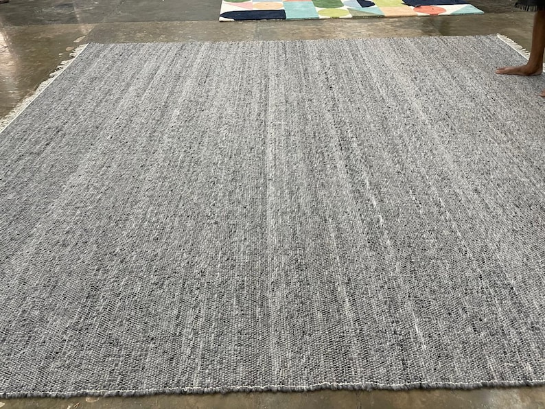 May include: A large, rectangular grey rug with a textured surface. The rug has a woven appearance with visible vertical lines and a fringe along the edge. The neutral colour and simple design make it suitable for various interior styles.