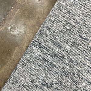 Könnte beinhalten: Nahaufnahme eines grau-weiß strukturierten Teppichs. Der Teppich hat ein gewebtes Muster und ist diagonal im Bildausschnitt angeordnet. Der Teppich befindet sich neben einer braunen Betonoberfläche.