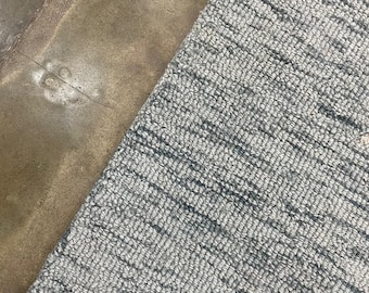 May include: A close-up of a gray and white textured rug. The rug has a woven pattern and is angled diagonally across the frame. The rug is next to a brown concrete surface.