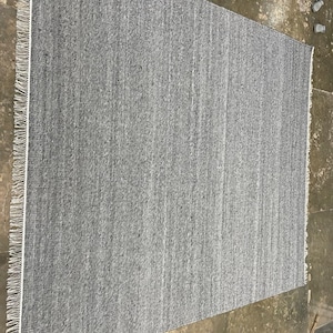 May include: A rectangular grey rug with a textured surface and a white fringe along one edge. The rug is made of woven material and has a simple, neutral design. The rug is suitable for various rooms.