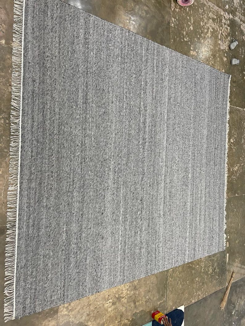 May include: A rectangular grey rug with a textured surface and a white fringe along one edge. The rug is made of woven material and has a simple, neutral design. The rug is suitable for various rooms.