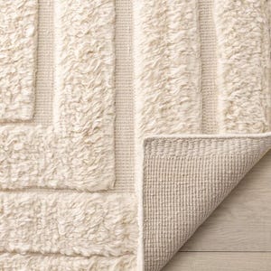Könnte beinhalten: Cremefarbener Teppich mit geometrischem Muster. Der Teppich hat ein strukturiertes, zotteliges Design mit erhabenen, rechteckigen Formen und einem gewebten Rand. Die Ecke des Teppichs ist umgeklappt und zeigt die Unterseite.