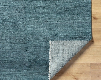 May include: A teal rug with a textured surface. The corner is folded back, showing the lighter backing. The rug is on a light brown wooden floor. The rug's dimensions are not visible.