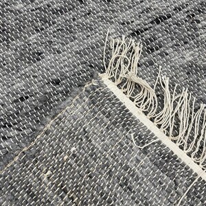 May include: Close-up of a grey and white woven rug. The rug has a textured appearance with a mix of grey and black threads. The edge of the rug is visible, showing a white border with fringe.