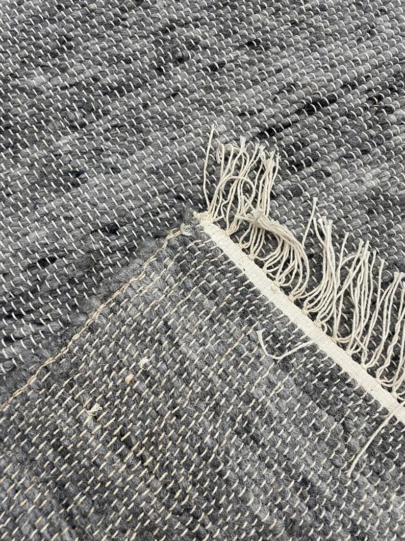 May include: Close-up of a grey and white woven rug. The rug has a textured appearance with a mix of grey and black threads. The edge of the rug is visible, showing a white border with fringe.