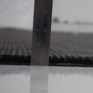 Może przedstawiać: Ciemnoszary, teksturowany dywan o niskim runie. Stalowa linijka jest ustawiona pionowo przy krawędzi dywanu, wskazując grubość około 0,6 cm. Dywan charakteryzuje się sp&oacute;jnym, liniowym wzorem w prążki na całej swojej powierzchni.