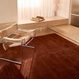 May include: A modern interior scene featuring a rust-colored rug with a geometric pattern. A beige chair with a metal frame sits on the left. A wooden tray with game pieces rests on a ledge, and a paper lantern hangs in the background.