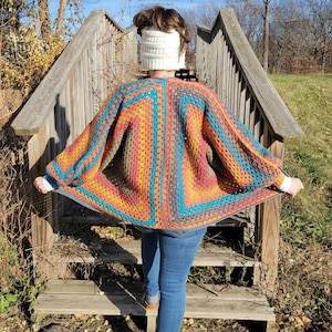 Könnte beinhalten: Ein farbenfroher, handgehäkelter Schal in Türkis, Orange, Gelb und Braun. Der Schal hat ein quadratisches Muster und wird über den Schultern getragen. Die Person trägt ein weißes Stirnband und blaue Jeans.