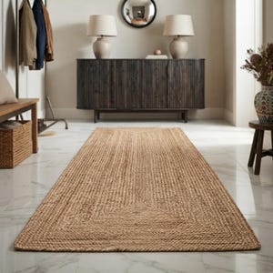 May include: A rectangular area rug crafted from natural tan woven fibers, featuring a distinct braided border design. The rug is positioned lengthwise on a white marble floor, extending from the foreground toward a dark wooden sideboard in the background.