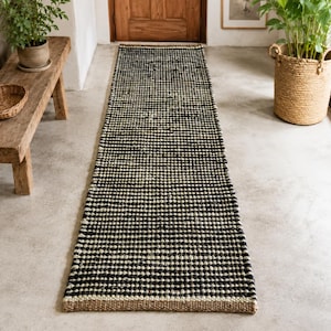Op de afbeelding: Een lange, rechthoekige loper met een geweven textuur en afwisselende horizontale strepen in zwart en gebroken wit. Het kleed heeft aan de onderkant een dikke rand van natuurlijk gekleurd jute of vezelmateriaal voor een contrast.
