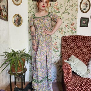 Può includere: Abito lungo con stampa floreale nei toni del giallo, rosa e verde. L'abito presenta maniche corte, scollatura quadrata e un bordo verde decorativo. L'abito è indossato con scarpe con plateau marroni.