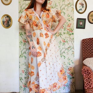 May include: A midi-length dress featuring a cream base with an orange and brown floral pattern. The garment has short flutter sleeves, a V-neckline trimmed with white lace, and a ruffled hemline. The fabric displays a mix of floral prints.