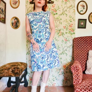 Peut inclure: Une robe sans manches arrivant au genou avec un motif floral cachemire bleu, bleu clair et blanc. La robe présente une encolure ronde, un corsage ajusté et une jupe droite, portée avec des bottines blanches.