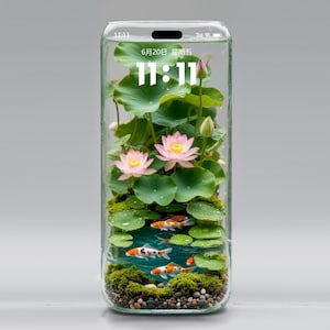 Könnte beinhalten: Ein gläsernes Terrarium in Form eines Telefons, das ein Miniatur-Aquasystem zeigt. Es zeigt rosa Lotusblumen, grüne Seerosenblätter und orange-weiß gestreifte Koi-Karpfen. Die Uhrzeit 11:11 wird angezeigt.