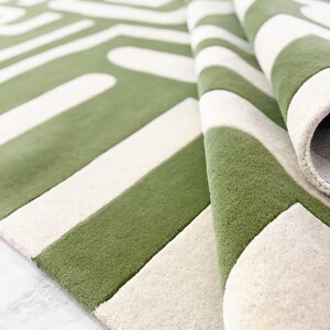 Puede incluir: Una alfombra de pelo denso con un patr&oacute;n geom&eacute;trico llamativo en verde oliva y blanco crema. La alfombra est&aacute; doblada para mostrar una textura suave y gruesa, junto con una base resistente. El dise&ntilde;o presenta l&iacute;neas anchas y rect&aacute;ngulos.