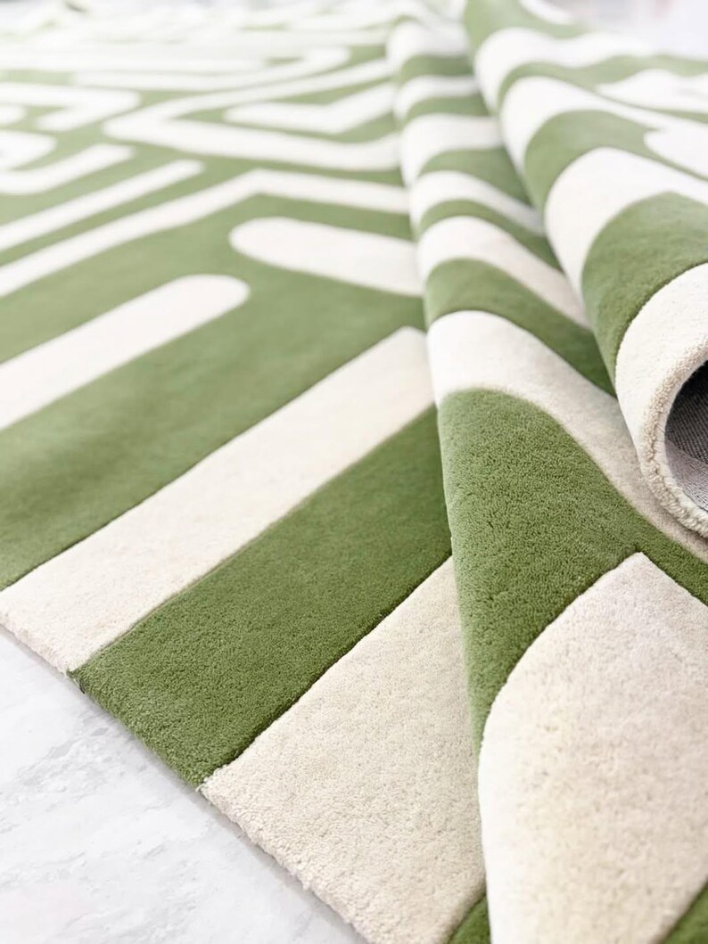 Puede incluir: Una alfombra de pelo denso con un patr&oacute;n geom&eacute;trico llamativo en verde oliva y blanco crema. La alfombra est&aacute; doblada para mostrar una textura suave y gruesa, junto con una base resistente. El dise&ntilde;o presenta l&iacute;neas anchas y rect&aacute;ngulos.