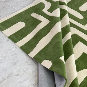 Puede incluir: Una alfombra rectangular con un patr&oacute;n geom&eacute;trico audaz en verde bosque profundo y blanco roto. La alfombra est&aacute; doblada para mostrar una superficie texturizada y copetuda en la parte superior y un reverso gris claro liso en la parte inferior.