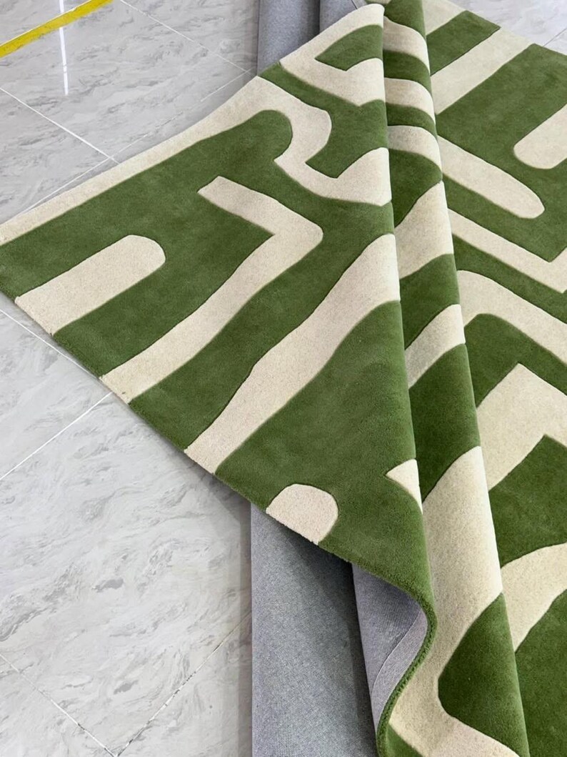 Puede incluir: Una alfombra rectangular con un patr&oacute;n geom&eacute;trico audaz en verde bosque profundo y blanco roto. La alfombra est&aacute; doblada para mostrar una superficie texturizada y copetuda en la parte superior y un reverso gris claro liso en la parte inferior.