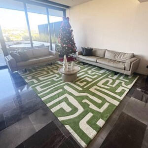 Puede incluir: Una alfombra grande con un patr&oacute;n geom&eacute;trico de laberinto en verde vibrante y crema. La alfombra est&aacute; situada en una habitaci&oacute;n con dos sof&aacute;s de cuero beige, una mesa de centro redonda con dos &aacute;rboles decorativos blancos y un &aacute;rbol de Navidad.