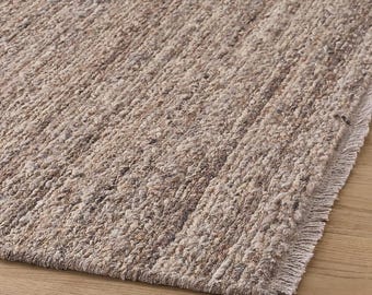 May include: A neutral-toned, textured rug with a woven appearance. The rug has a fringe detail on one edge and is made of a natural fiber. The color palette includes shades of beige and brown, creating a warm and inviting aesthetic.