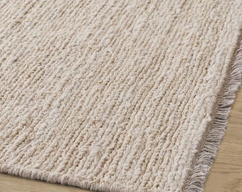 May include: Close-up of a beige textured rug with a fringe edge. The rug has a woven, ribbed pattern, and the neutral color would complement various interior design styles. The rug is placed on a light-colored wooden floor.