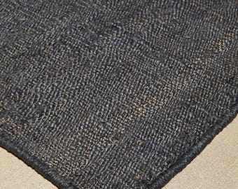 May include: A close-up of a dark grey woven rug with a textured surface. The rug has a dark border and a neutral background. The rug appears to be made of natural fibres.