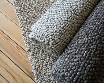 May include: Three rolled textured rugs in shades of gray and beige rest on a wooden floor. The rugs have a nubby, bubble-like texture. The colors range from light beige to dark gray.
