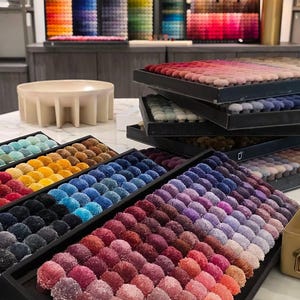 Puede incluir: Varias bandejas de exhibici&oacute;n rectangulares que contienen filas de peque&ntilde;os pompones de tela redondos y texturizados en un amplio espectro de colores, incluyendo tonos de rojo, rosa, morado, azul, amarillo y marr&oacute;n, dispuestos en degradados.