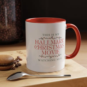 May include: White ceramic mug with a red interior and handle. The mug features the text "THIS IS MY HALLMARK CHRISTMAS MOVIE WATCHING MUG" in red. A spoon and a macaron are on a wooden surface.