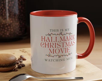 May include: White ceramic mug with a red interior and handle. The mug features the text "THIS IS MY HALLMARK CHRISTMAS MOVIE WATCHING MUG" in red. A spoon and a macaron are on a wooden surface.