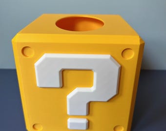 May include: A yellow cube-shaped box with a white question mark on one side, reminiscent of a video game. The box has a circular opening on top and circular details on the sides. The design is a vibrant yellow and white.