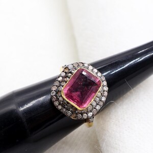 Op de afbeelding: Een gouden ring met een grote, rechthoekige, roze edelsteen in het midden, omringd door een halo van kleinere, heldere stenen. De ring wordt gepresenteerd op een zwarte standaard tegen een witte achtergrond.