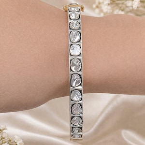 Könnte beinhalten: Ein silbernes Armband mit einer Reihe klarer, unregelmäßig geformter Steine, die in quadratischen Rahmen gefasst sind. Das Armband hat einen goldenen Verschluss und wird an einem Handgelenk präsentiert. Der Hintergrund ist ein weicher, weißer Stoff.