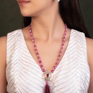 Puede incluir: Un conjunto de collar y pendientes a juego con un colgante central con una piedra preciosa roja rodeada de piedras blancas. El collar tiene una larga hebra de cuentas rosas con una borla. Los pendientes tienen forma de flor con una piedra roja en el centro.