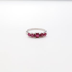 May include: A silver ring with five oval, vivid red gemstones. The gemstones are set in a row. The ring is shown against a white backdrop, accentuating the jewellery's design and colour. The ring is a classic piece of jewellery.
