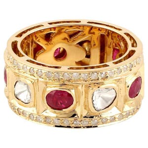 Può includere: Un anello d'oro con una fascia con pietre preziose rosse e chiare alternate. L'anello è incastonato con piccole pietre preziose chiare attorno alla fascia. L'interno dell'anello ha un design decorativo e aperto.