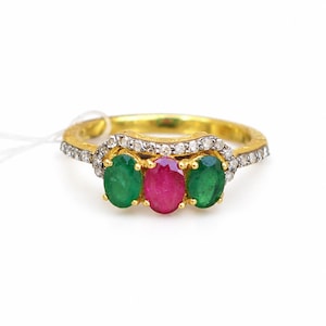 Puede incluir: Un anillo de oro con un rubí central de corte ovalado flanqueado por dos esmeraldas de corte ovalado. El engaste incluye una fila curva de pequeñas piedras blancas redondas sobre las gemas, con piedras blancas adicionales en el aro.