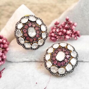Puede incluir: Dos pendientes de botón con forma floral. Cada pendiente presenta un grupo central de pequeñas piedras preciosas redondas de color rojo oscuro, rodeadas de piedras preciosas claras, más grandes y de forma irregular. Los pendientes tienen un engaste de metal oscuro y una base dorada.